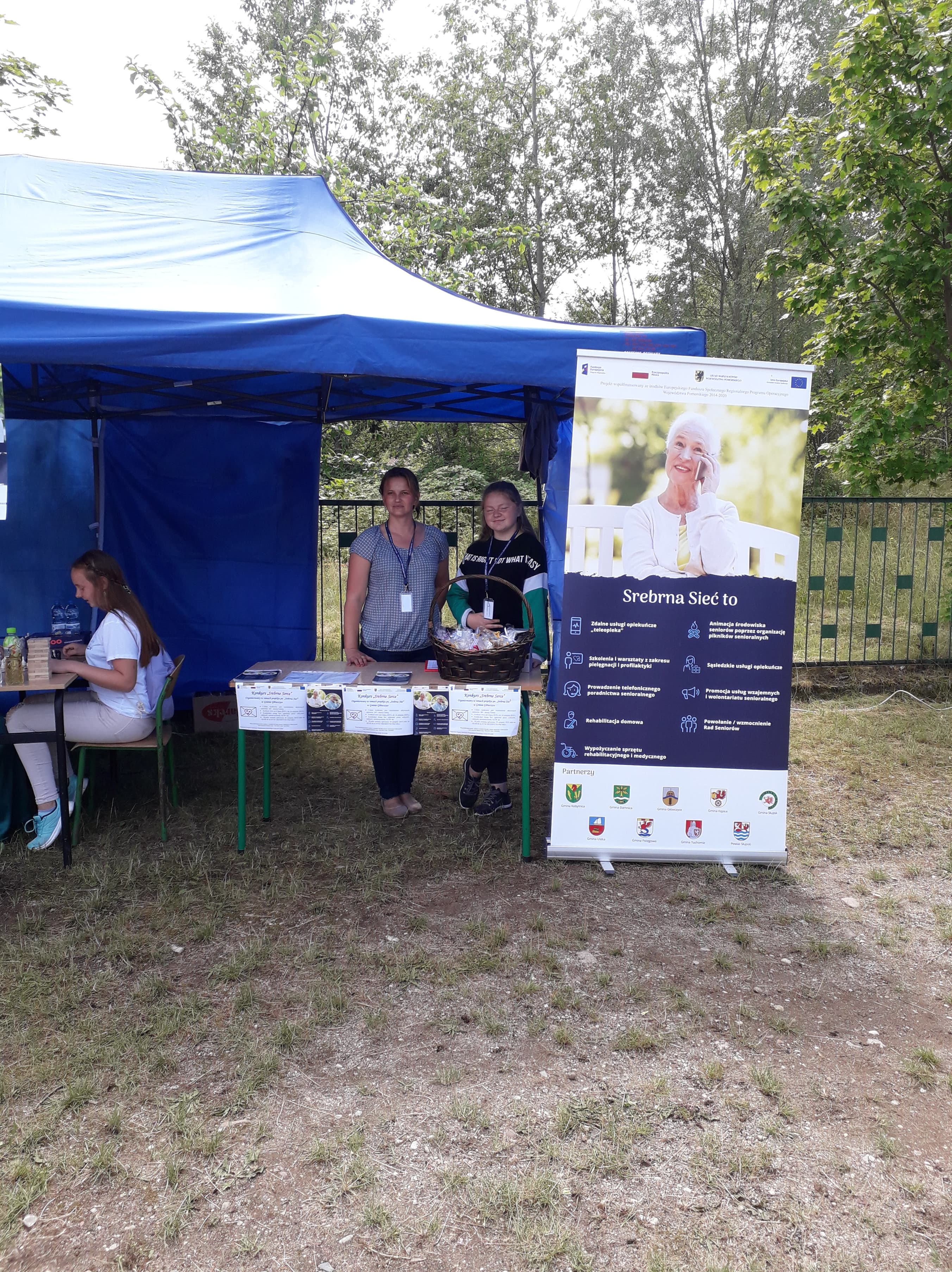 Wolontariusz wraz z opiekunem wolontariatu pod namiotem z grami planszowymi i karcianymi, z koszem łakoci dla uczestników festiwalu oraz plakatami i ulotkami dotyczącymi projektu Srebrna Sieć, w tym organizowanego konkursu pn. Srebrne Serce.