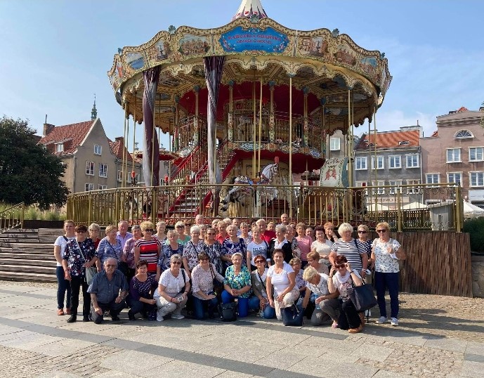 Liczna grupa uczestników wycieczki na tle kolorowej karuzeli gdańskiej.