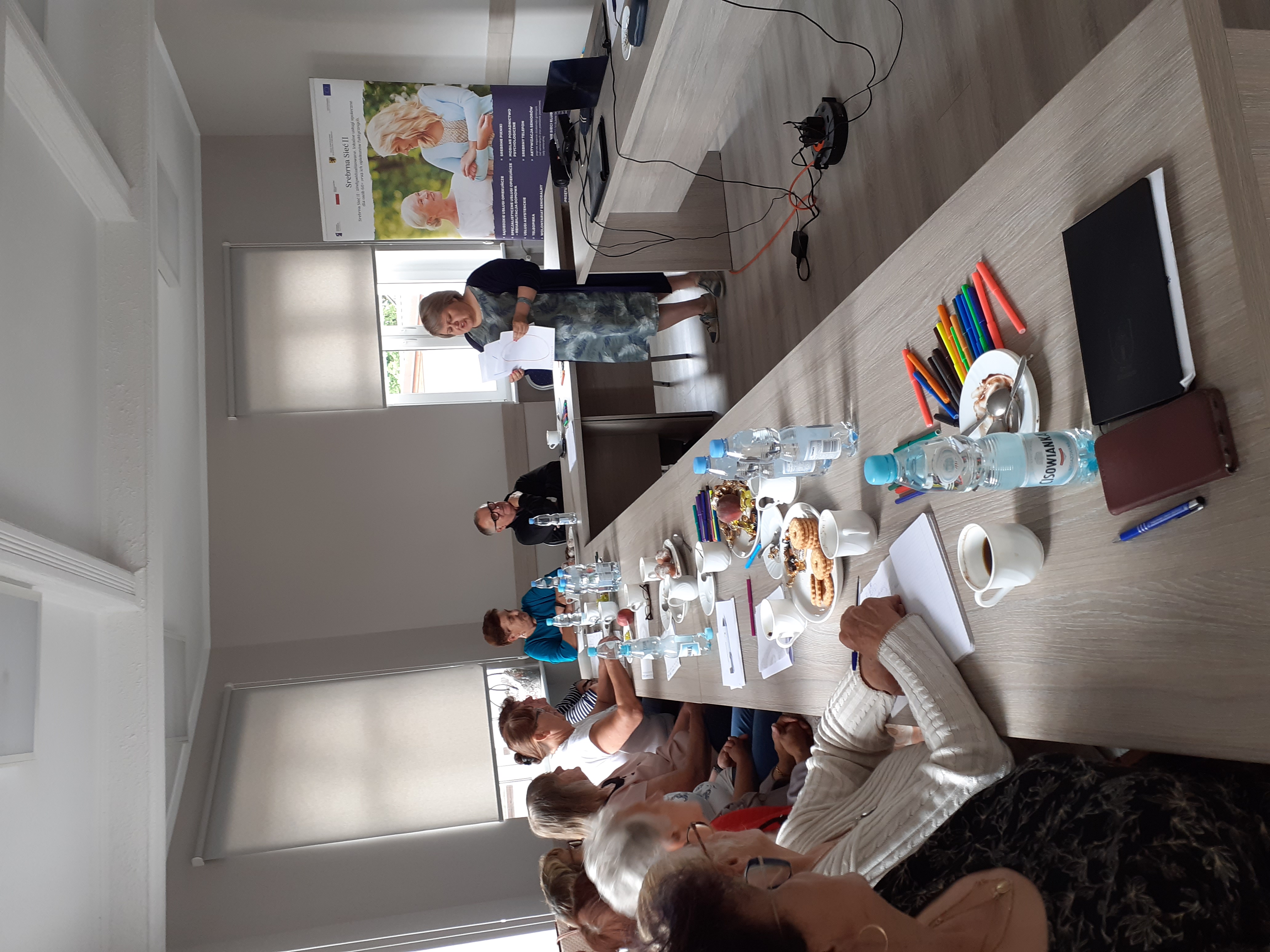 Uczestnicy szkolenia siedzący przy stole i pani szkoląca stojąca przed nimi