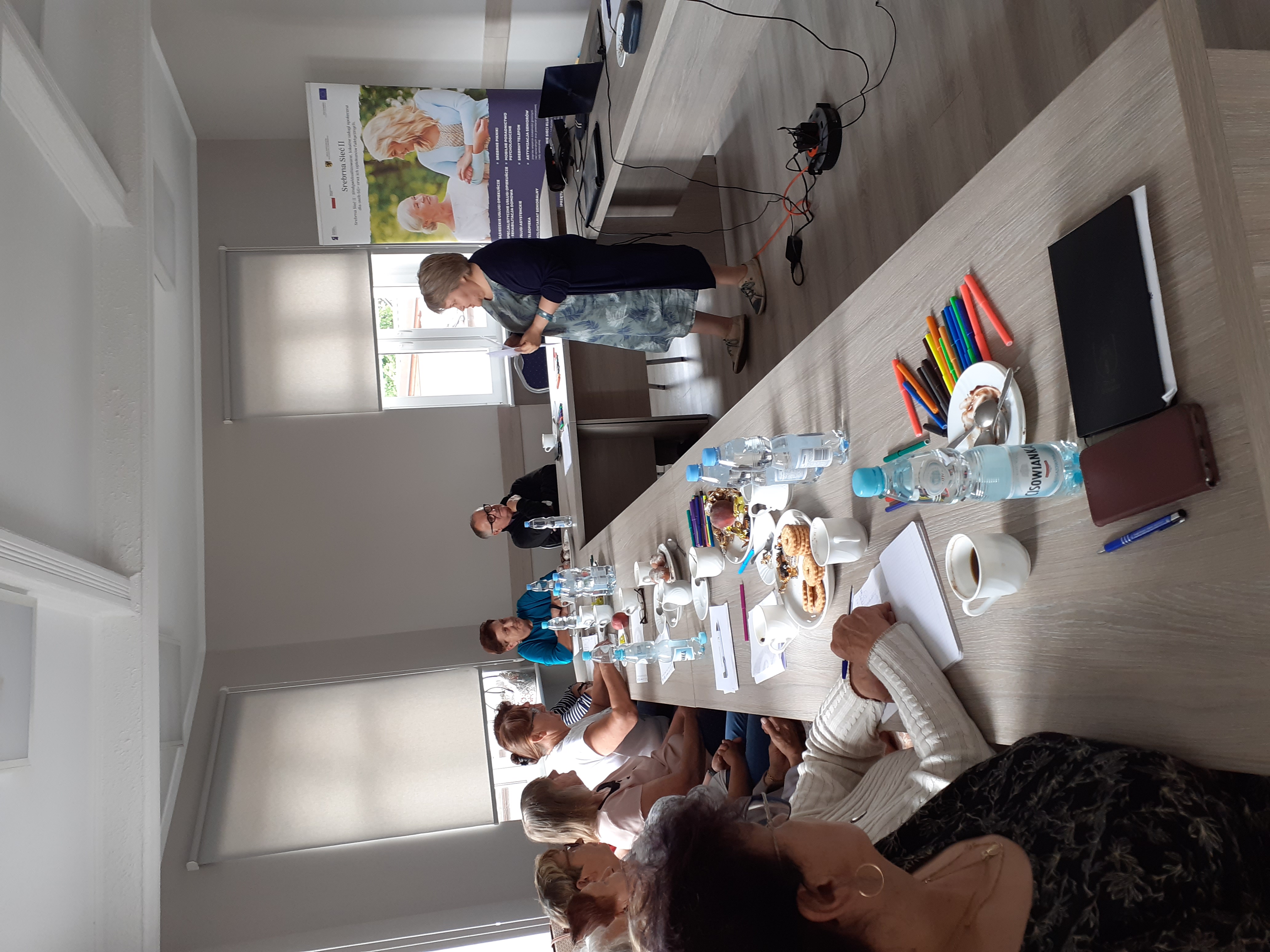 Uczestnicy szkolenia siedzący przy stole i pani szkoląca stojąca przed nimi